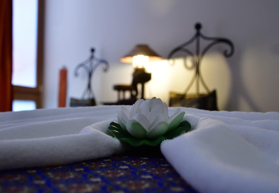 Uma sala de massagens tailandesas no salão Nattakan Thai Massage, com uma toalha branca cuidadosamente dobrada e uma flor de lótus branca artificial no centro. O fundo está suavemente iluminado, realçando uma atmosfera serena e calmante com uma visão desfocada do mobiliário ornamentado, incluindo um candeeiro e cadeiras decorativas.