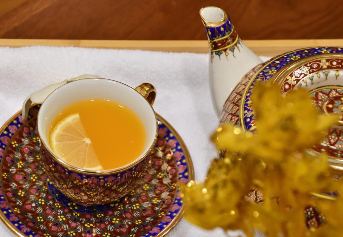 Ornate ceramic teacup and saucer filled with orange tea and a slice of lemon, accompanied by a matching teapot, placed on a white towel. The intricate design features rich blue and gold patterns, adding a touch of elegance.