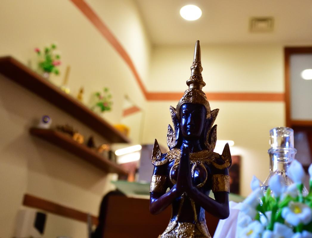 A serene sculpture of a traditional Thai deity with hands in a prayer position is displayed in the foreground of a warmly lit room. The background features wall shelves with small decorative items and potted plants, enhancing the calming ambiance of the Nattakan Thai Massage salon.