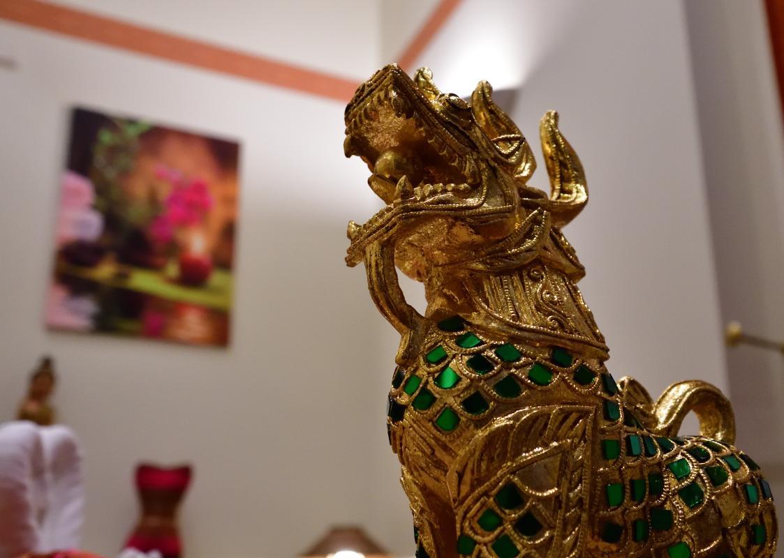 Golden Thai dragon statue with intricate designs, featuring green accents. In the background, an out-of-focus painting with pink and red flowers, creating a relaxing ambiance inside Nattakan Thai Massage salon.