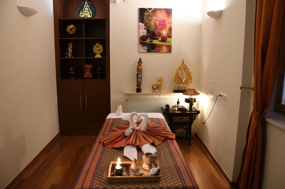 A serene massage room at Nattakan Thai Massage salon, featuring a neatly made massage bed adorned with intricately folded towels, golden accents, and candles. The warm lighting and wooden floor create a peaceful ambiance. The room is decorated with traditional Thai artifacts, including a wooden shelf with golden ornaments, statues, and a framed floral artwork on the wall.