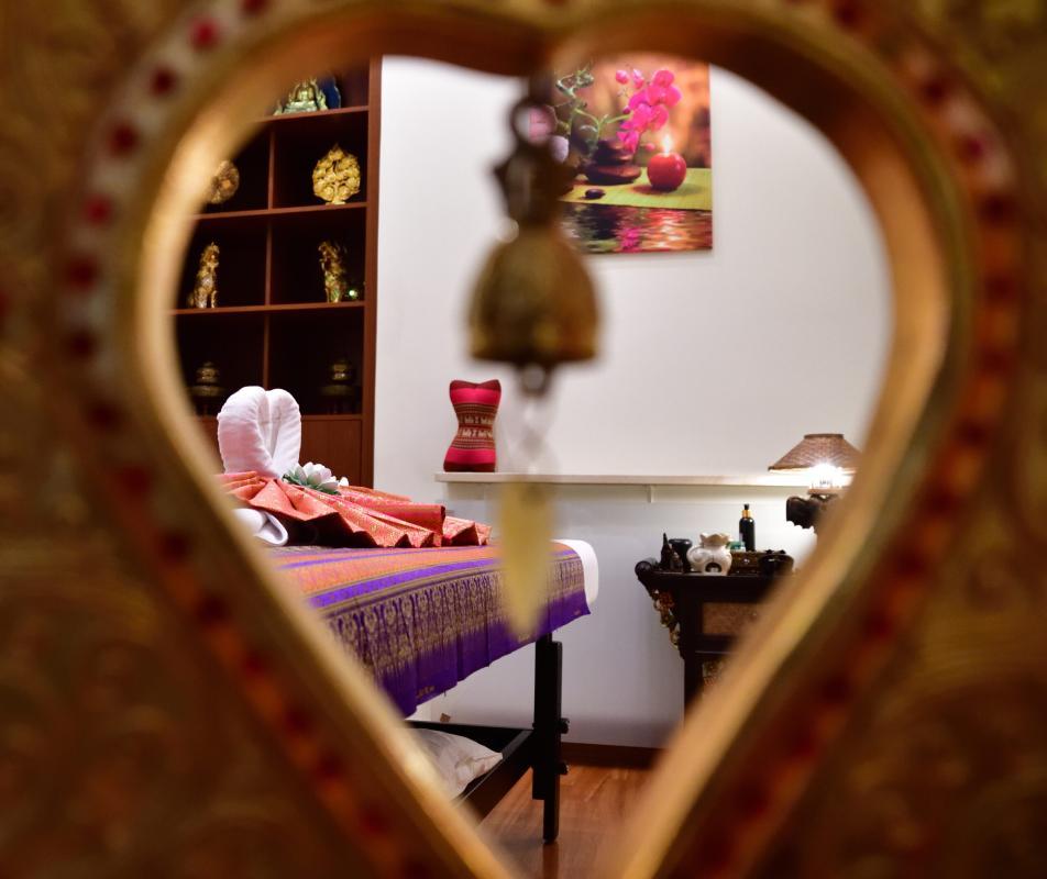 Nhìn qua đồ trang trí hình trái tim tập trung vào phòng mát-xa nhẹ nhàng tại Nattakan Thai Massage. Phòng có giường mát-xa với khăn gấp và lối trang trí truyền thống của Thái Lan. Phía sau là kệ trưng bày các bức tượng vàng và bức tranh vẽ hoa lan.