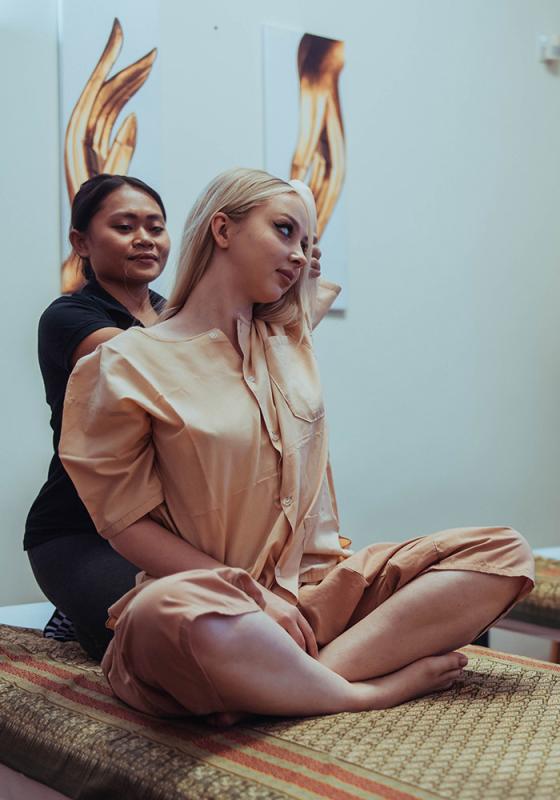 A massage therapist at Nattakan Thai Massage performs a traditional Thai massage on a client dressed in comfortable attire, gently stretching her upper body. The setting is serene with soft lighting and artistic wall decorations in the background.