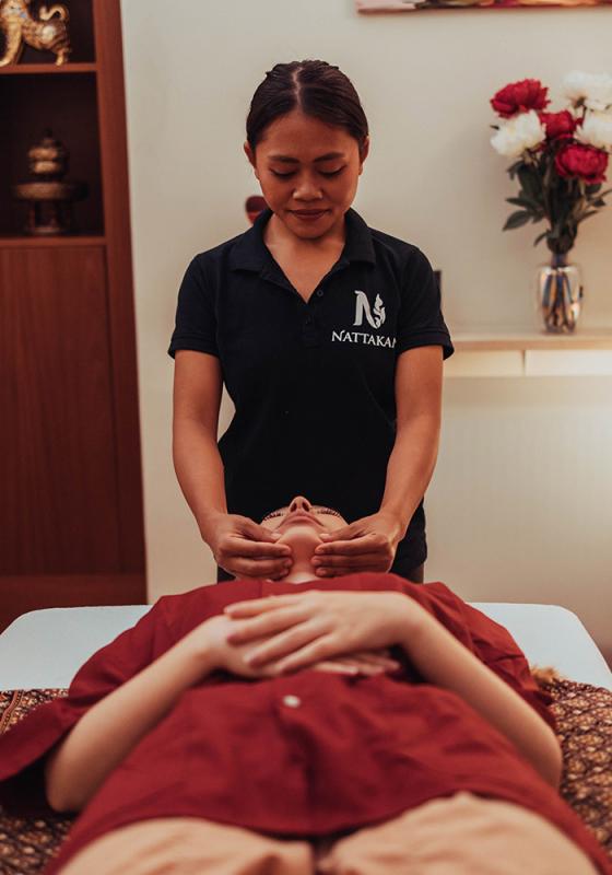 Une masseuse expérimentée, vêtue d'une chemise noire portant le logo Nattakan, effectue un massage sur un client détendu, allongé dans une pièce paisible ornée de fleurs et d'un décor traditionnel.