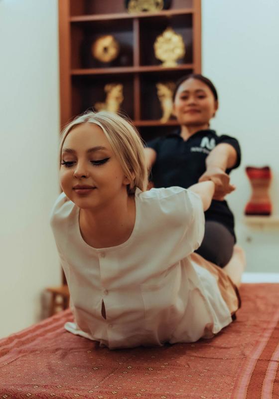 Mujer sonriente recibiendo un masaje tradicional tailandés de espalda en el salón de masajes tailandés Nattakan. El terapeuta tira de sus brazos hacia atrás mientras ella se estira en una camilla de masaje, con un ambiente sereno creado por objetos decorativos dorados de fondo.