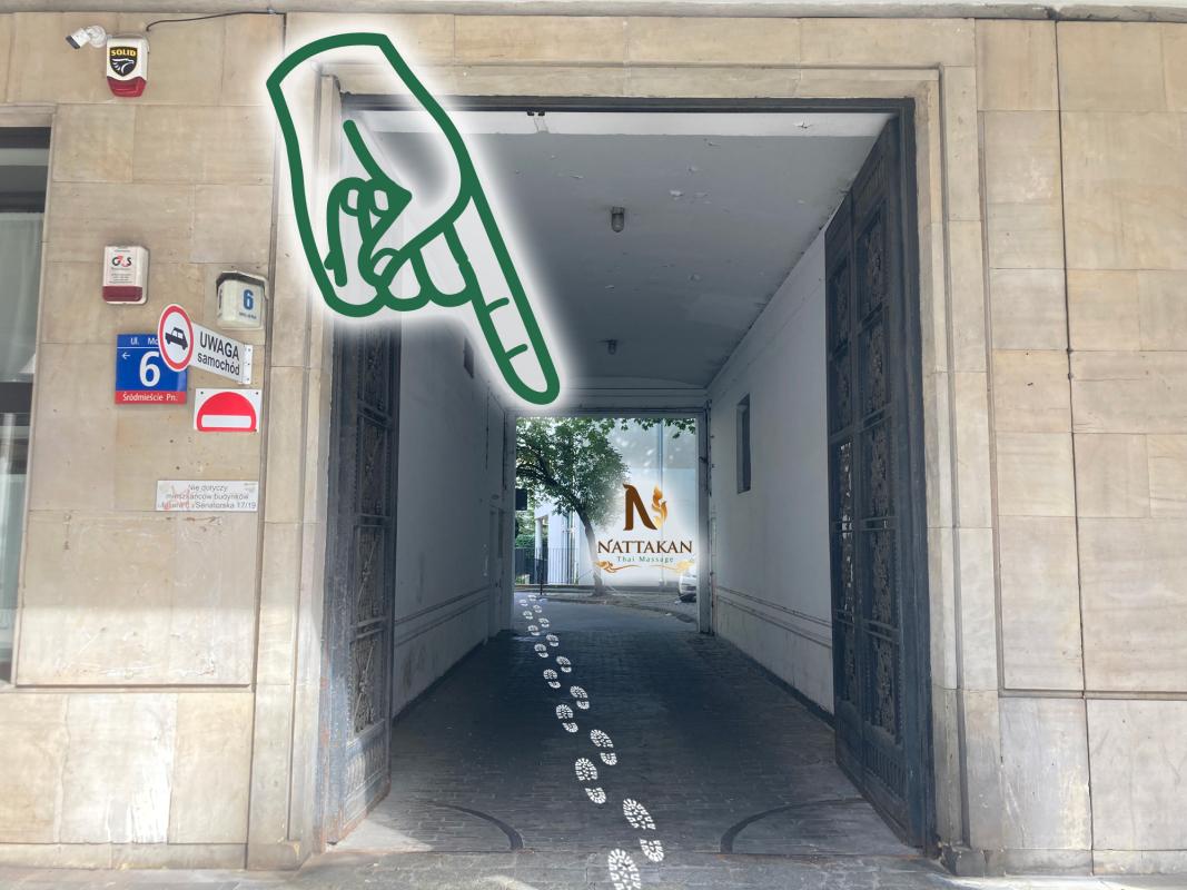 Ingresso di un edificio con una grande mano grafica che indica il corridoio interno. Le impronte sul terreno conducono a una porta di vetro con il logo Nattakan Thai Massage visibile sullo sfondo. Molteplici insegne sono attaccate al muro sul lato sinistro dell'ingresso.