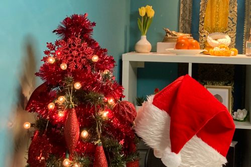 Festive scene within a Thai massage salon featuring a small, decorated Christmas tree adorned with red ornaments and lights. A Santa hat is placed on the corner of a cushioned massage table, and a shelf in the background displays a vase with yellow tulips and traditional Thai decor.