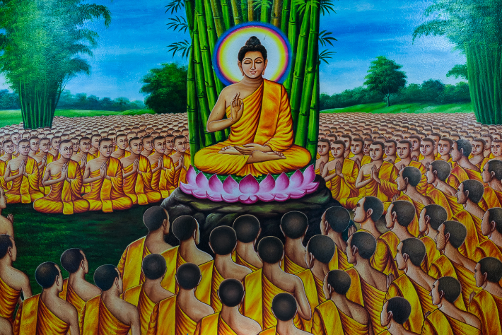 Une peinture vibrante représente Bouddha méditant assis sur une plate-forme de fleurs de lotus, entouré de nombreux moines vêtus de robes jaunes. Ils écoutent attentivement le sermon de Bouddha dans un cadre extérieur luxuriant composé de champs verts et de grands bambous sous un ciel lumineux.