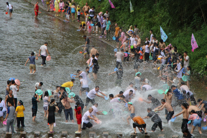 色とりどりのバケツで水を掛け合い、浅瀬の川で水合戦を楽しむ大勢の人々。川岸と水中の参加者が戯れに夢中になっており、それを横から見ている観客もいる。背景には緑が見え、賑やかで活気のある光景である。