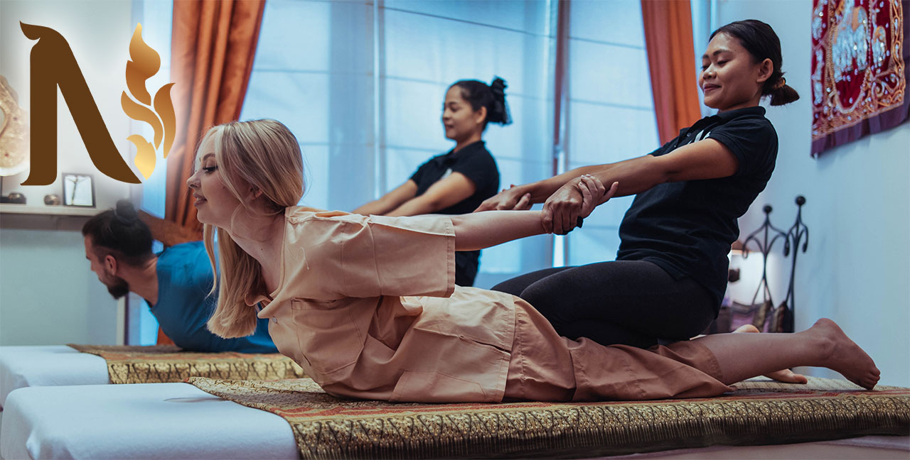 Une femme en tenue de massage beige reçoit un massage thaïlandais traditionnel d'un thérapeute qui lui étire doucement le dos en tirant sur ses bras. Une autre personne reçoit un traitement similaire à l'arrière-plan. L'éclairage et le décor sont chaleureux et le logo N de Nattakan Thai Massage se trouve au premier plan.