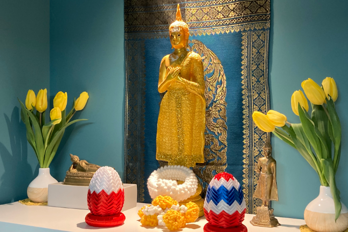 En gyllen Buddhastatue prydet med et blått og gyllent billedvev i bakgrunnen, omgitt av gule tulipaner i hvite vaser. To fargerike, intrikat utformede eggformede dekorasjoner og tradisjonelle thailandske elementer er plassert på det hvite alteret.