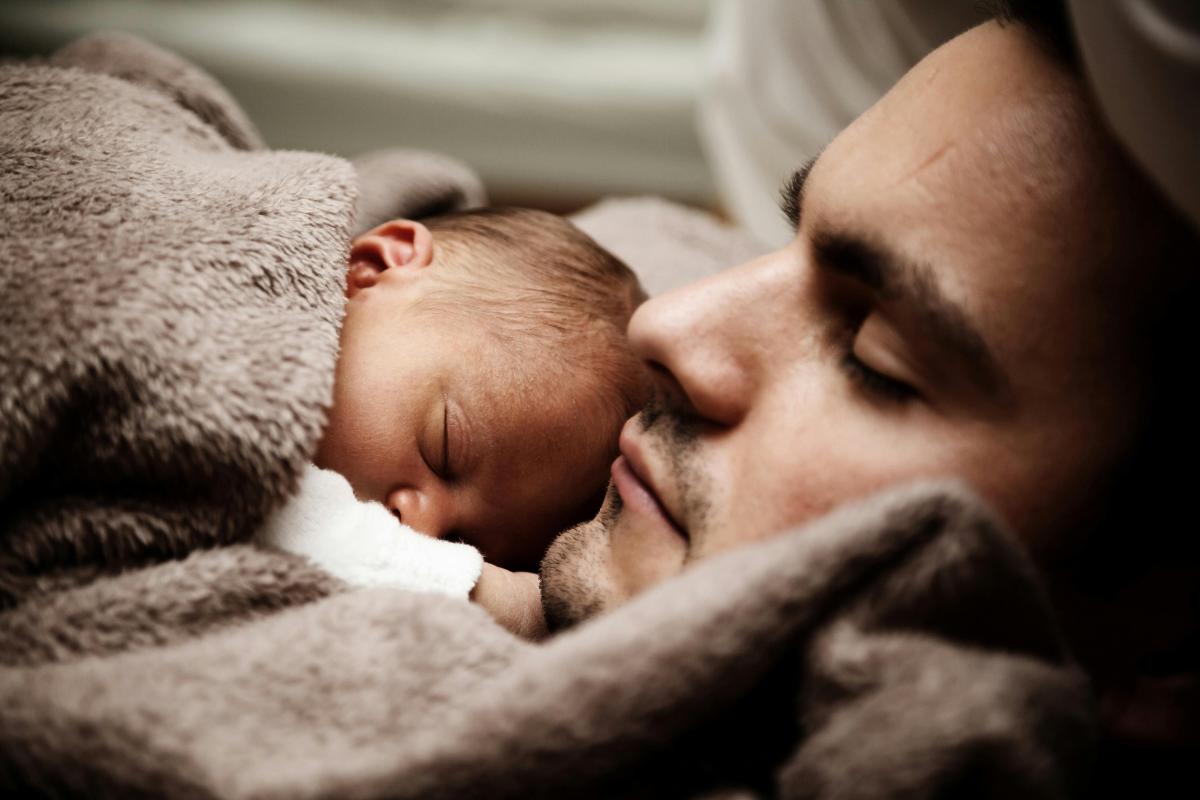 A close-up of a person and a baby sleeping together under a soft, beige blanket. The babys face is nestled close to the persons face, both appearing peaceful and content.