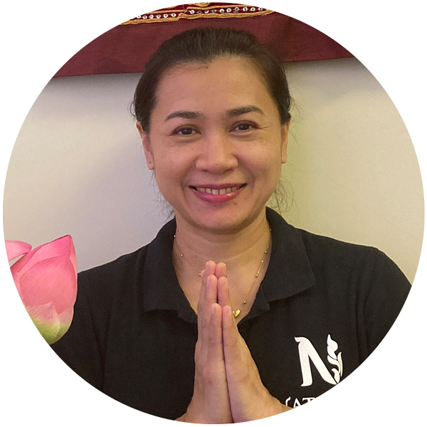 Một người mỉm cười mặc áo polo đen có logo Nattakan Thai Massage ở bên trái, chắp hai tay vào nhau theo cử chỉ chào truyền thống của Thái Lan. Một bông hoa sen màu hồng hiện rõ ở phía bên trái.