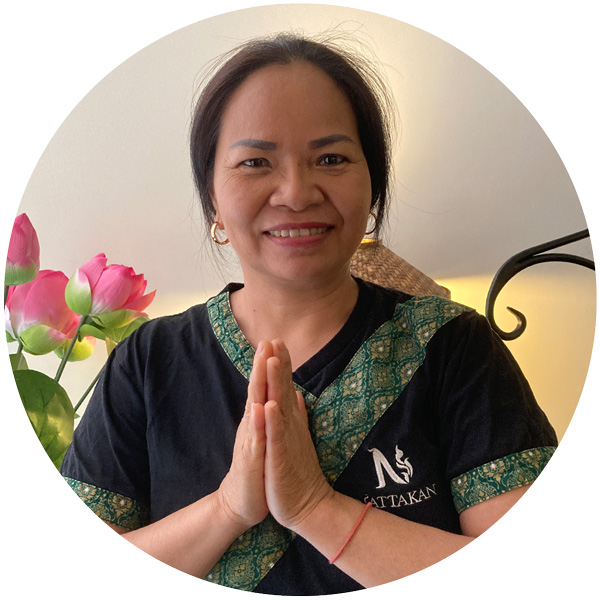 A woman wearing a black shirt with green decorative trim and a logo that says Nattakan stands with her hands pressed together in a traditional Thai greeting gesture. She is smiling warmly, and there are pink flowers in the background.