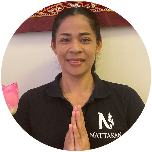 Smiling massage therapist in a black shirt with the Nattakan Thai Massage logo, holding hands in a traditional Thai greeting.