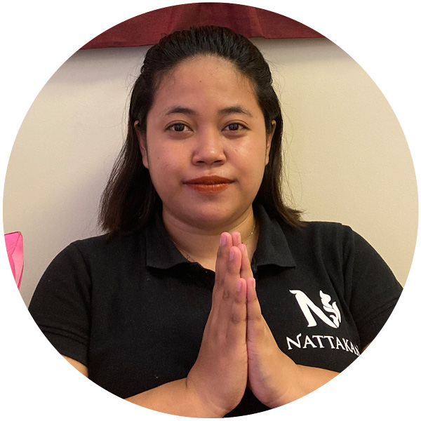 A massage therapist at Nattakan Thai Massage wearing a black polo shirt with the salons logo, holding hands together in a traditional Thai greeting (wai).